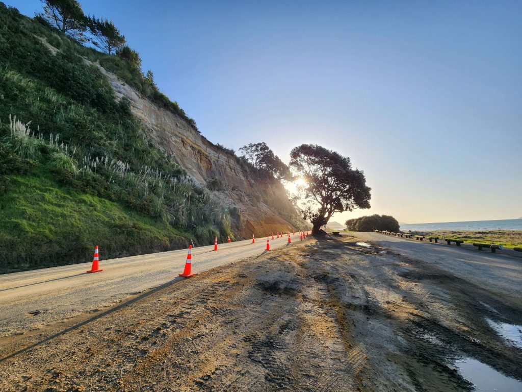 Roadworks along the coast with a Pohutukawa tree visible in front of a sunset.